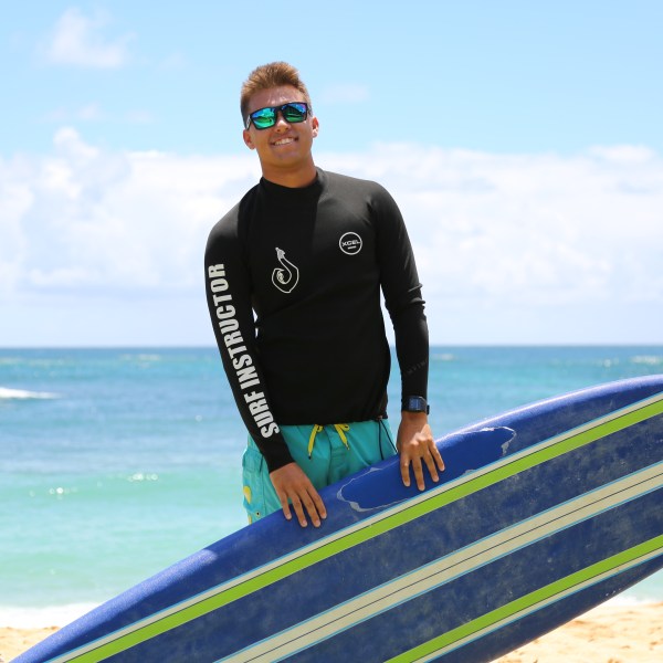 man in a surf instructor rash guard holding his surfboard at his feet with the ocean in the background