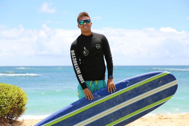 man in a surf instructor rash guard holding his surfboard at his feet with the ocean in the background