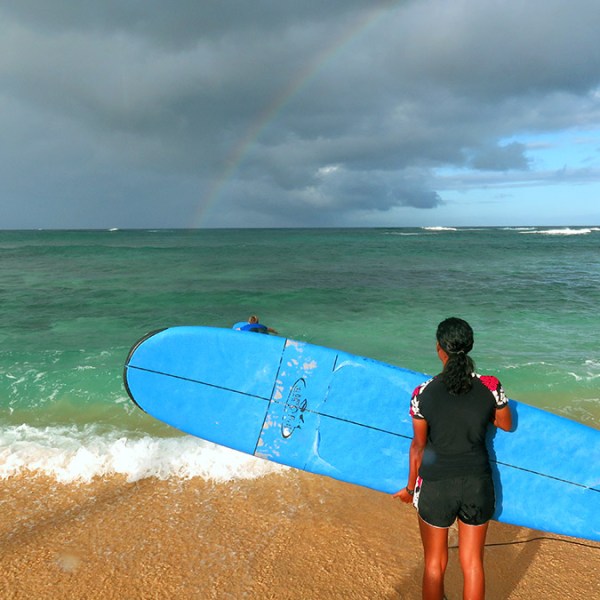girl holding her surfboard sideways looking at the storm clouds and rainbow