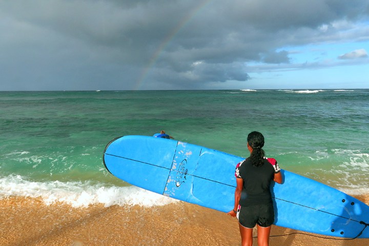 girl holding her surfboard sideways looking at the storm clouds and rainbow