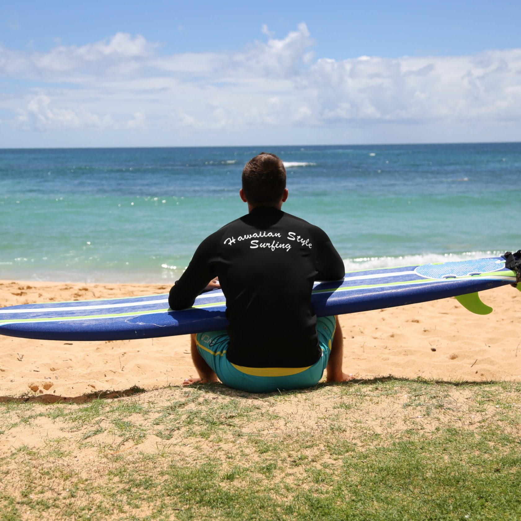 a man sitting with his back to us overlooking the ocean holding his surfboard while he waxes it and contemplates
