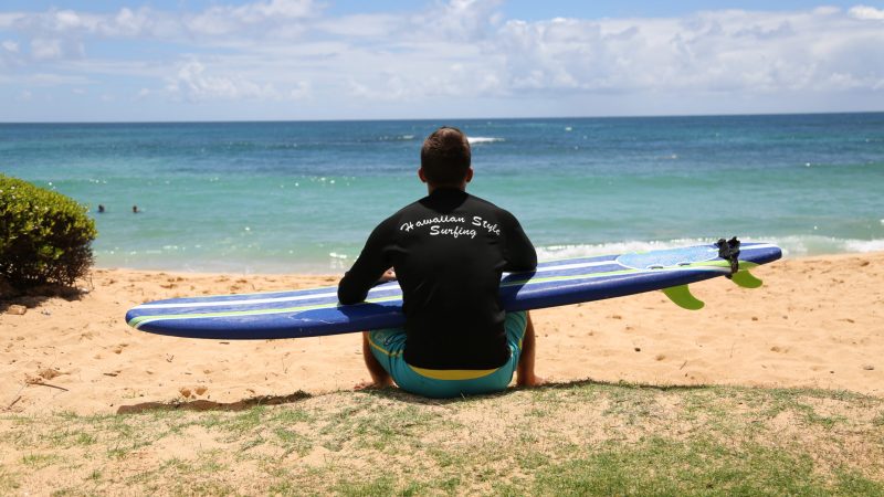 a man sitting with his back to us overlooking the ocean holding his surfboard while he waxes it and contemplates
